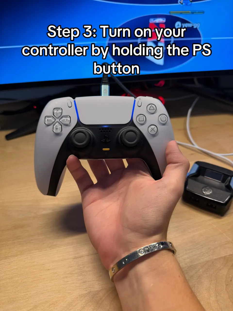 Hand holding a DualSense controller, thumb on the PS button, light bar lit